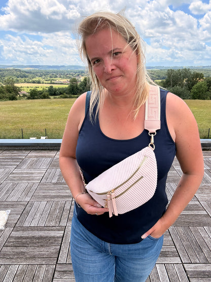 Sac banane velours côtelé rose pâle porté en bandoulière par une femme en débardeur bleu, avec deux poches zippées et sangle réglable.