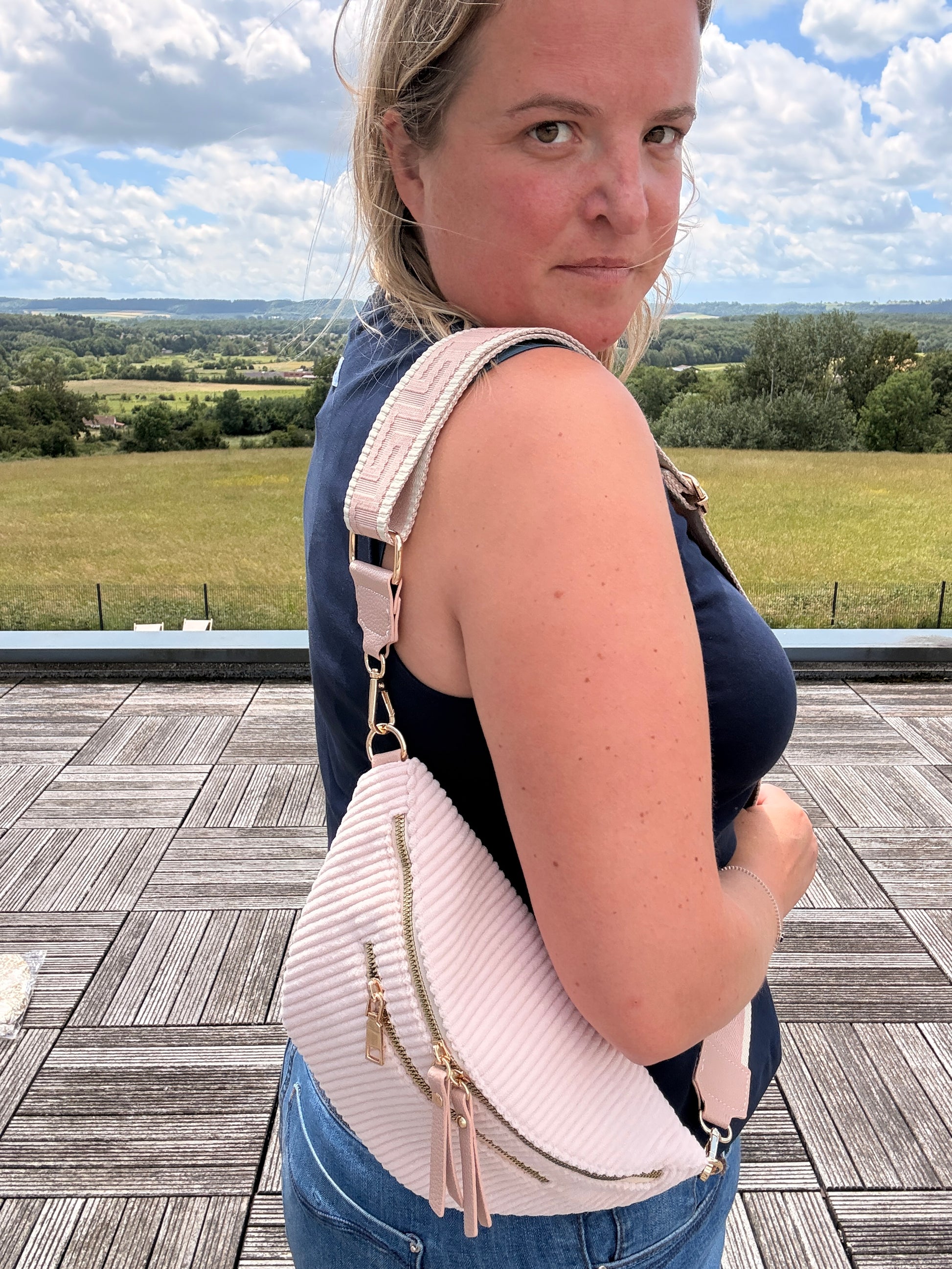 Sac banane velours côtelé rose pâle porté en bandoulière par une femme sur une terrasse, avec 2 poches zippées et sangle réglable.