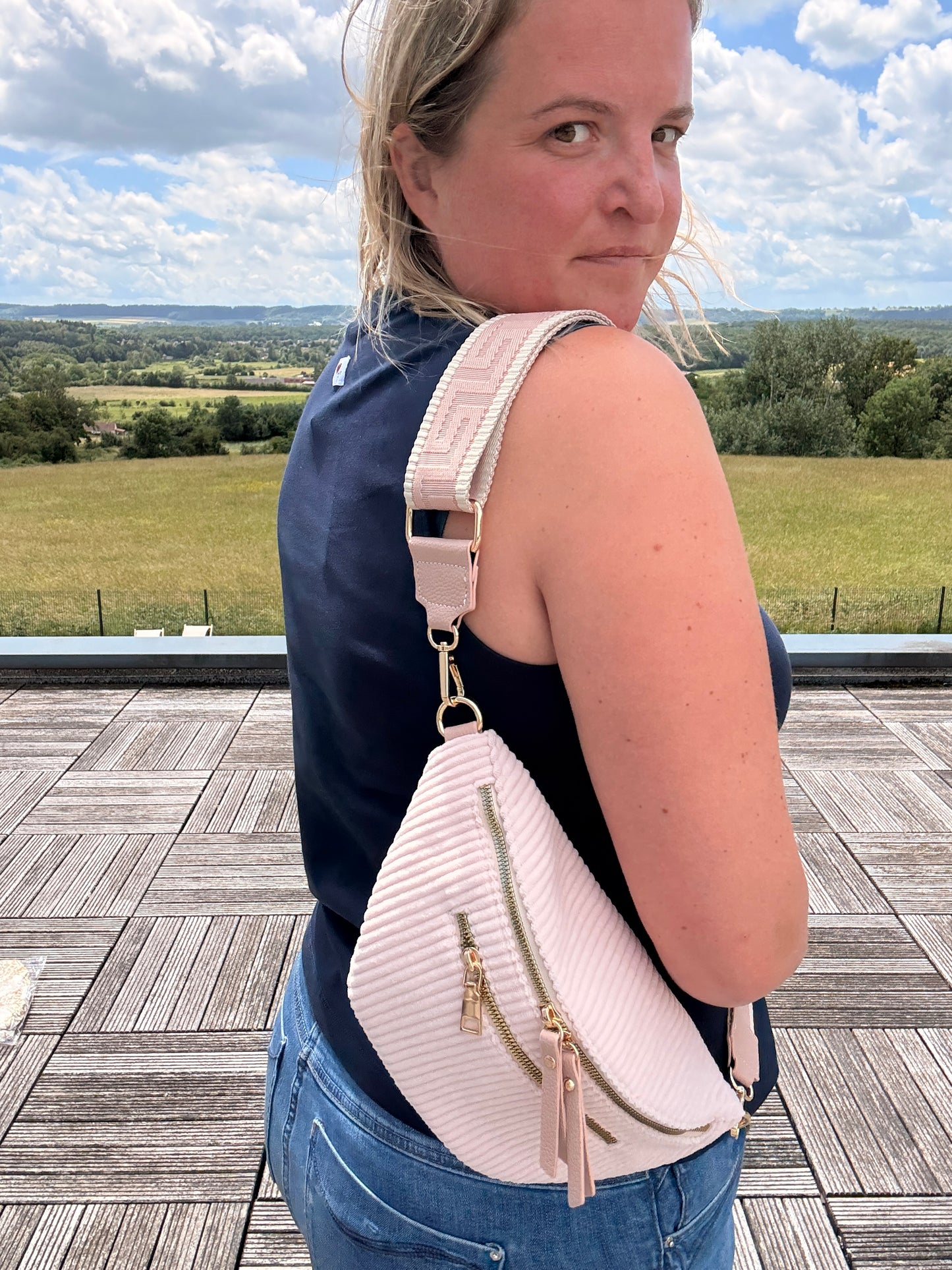 Sac banane velours côtelé femme en rose pâle porté en bandoulière, avec 2 poches zippées et sangle réglable visibles sur le côté.
