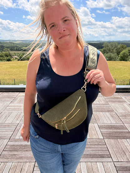 Sac banane velours côtelé kaki avec poches zippées portée en bandoulière par une femme, fond paysage et ciel bleu.