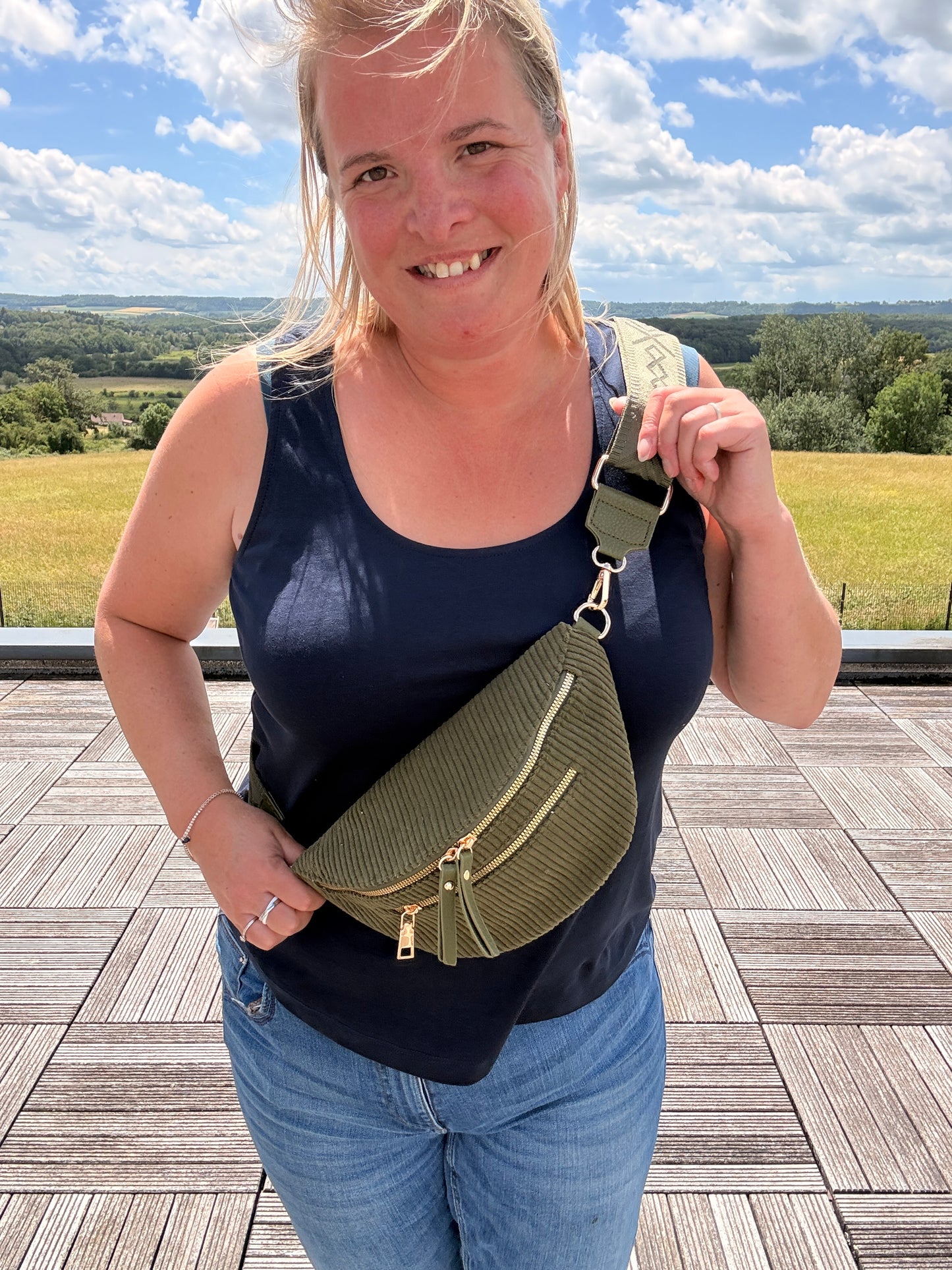 Femme portant un sac banane velours côtelé kaki avec poches zippées, debout en extérieur, sourire, horizon champ et ciel bleu en arrière-plan.