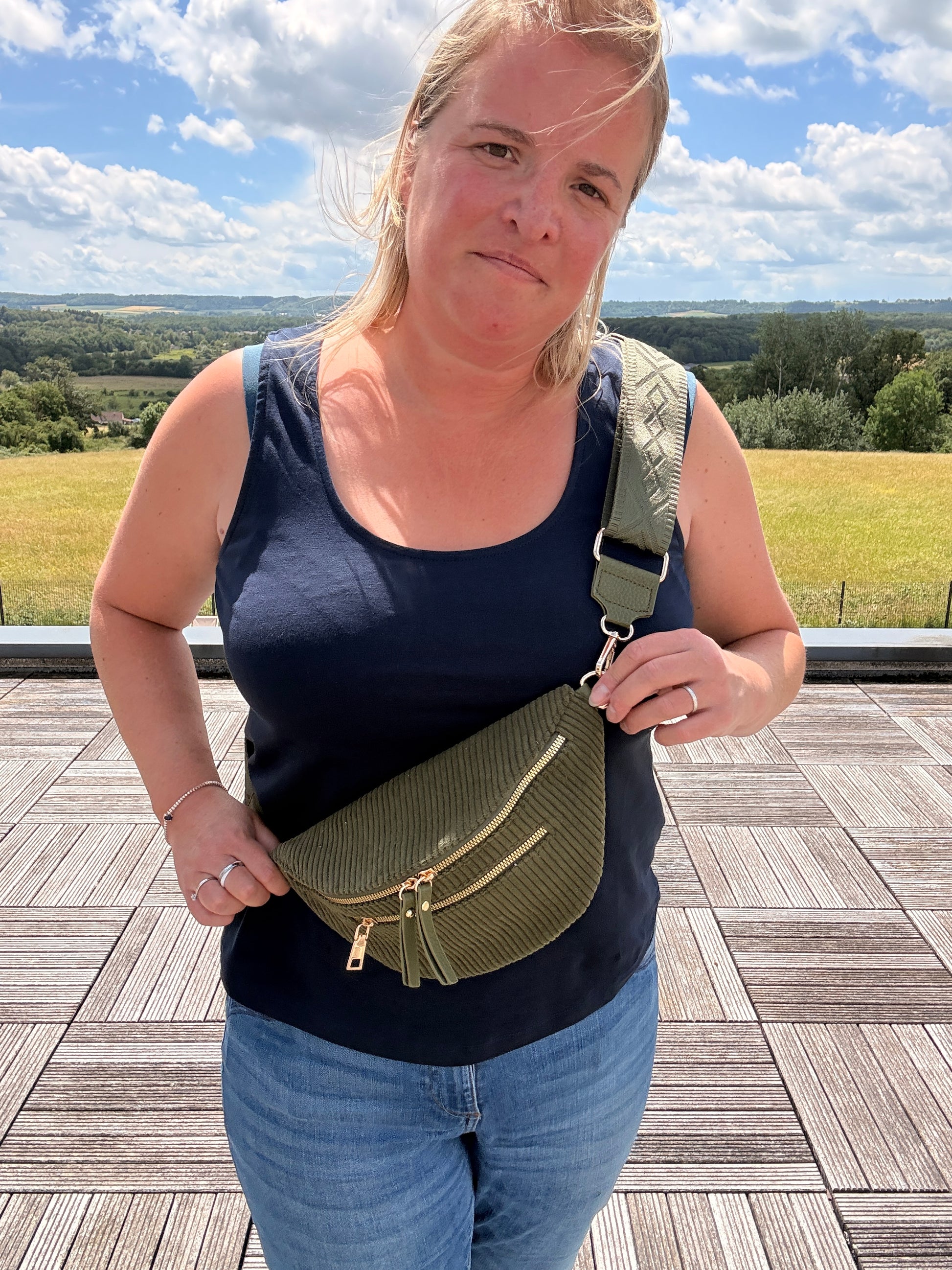 Sac banane velours côtelé kaki avec poches zippées porté en bandoulière sur une femme, horizon champêtre en arrière-plan.