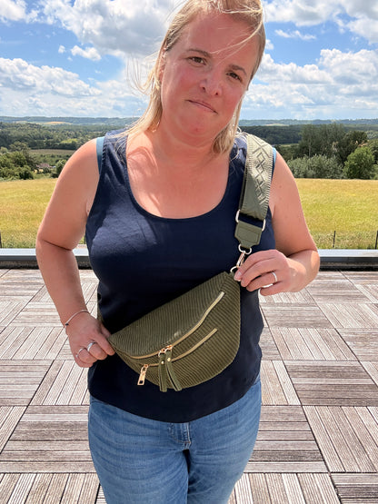 Sac banane velours côtelé kaki avec poches zippées porté en bandoulière sur une femme, horizon champêtre en arrière-plan.