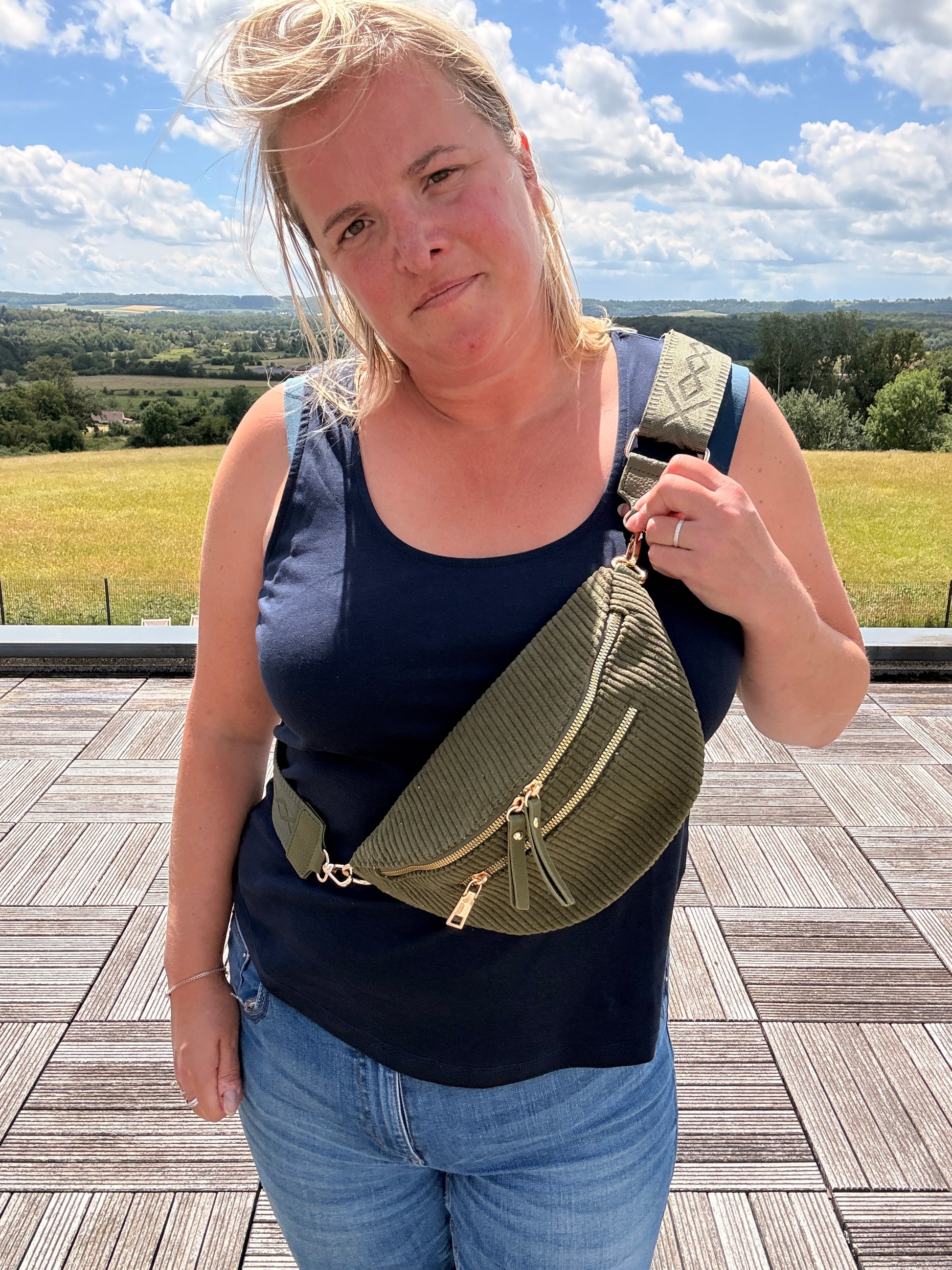 Sac banane velours côtelé kaki avec poches zippées porté sur l’épaule d’une femme, en plein air, paysage champêtre à l’horizon.