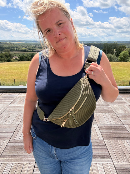 Sac banane velours côtelé kaki avec poches zippées porté sur l’épaule d’une femme, en plein air, paysage champêtre à l’horizon.