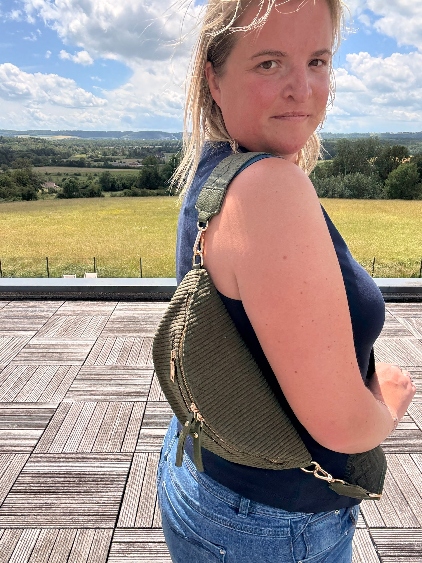 Sac banane velours côtelé kaki porté en bandoulière par une femme, vue de profil en extérieur avec paysage Champ clair et ciel bleu. Focus sur les poches zippées et la texture côtelée de la sacoche.
