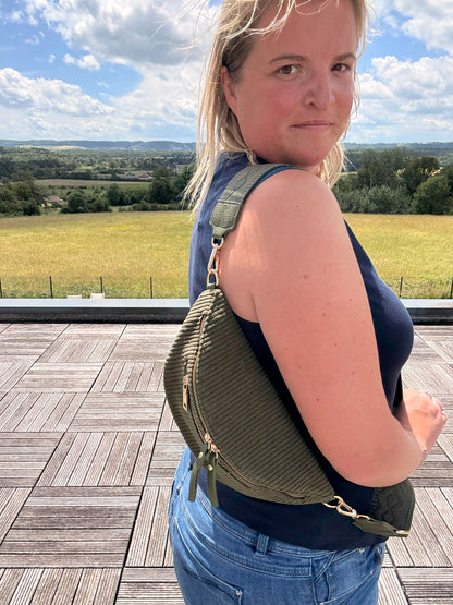 Sac banane velours côtelé kaki porté en bandoulière par une femme, vue de profil en extérieur avec paysage Champ clair et ciel bleu. Focus sur les poches zippées et la texture côtelée de la sacoche.