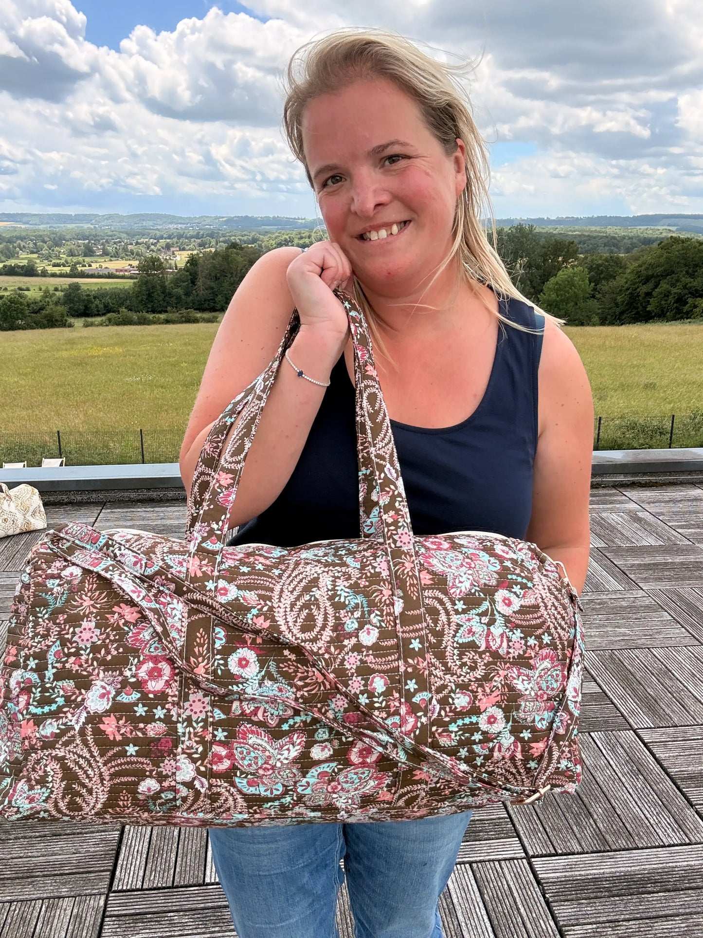 Sac polochon matelassé fleuri paisley porté par une femme sur un pont, prêt à voyager, bandoulière incluse. Grand sac de voyage bohème chic.