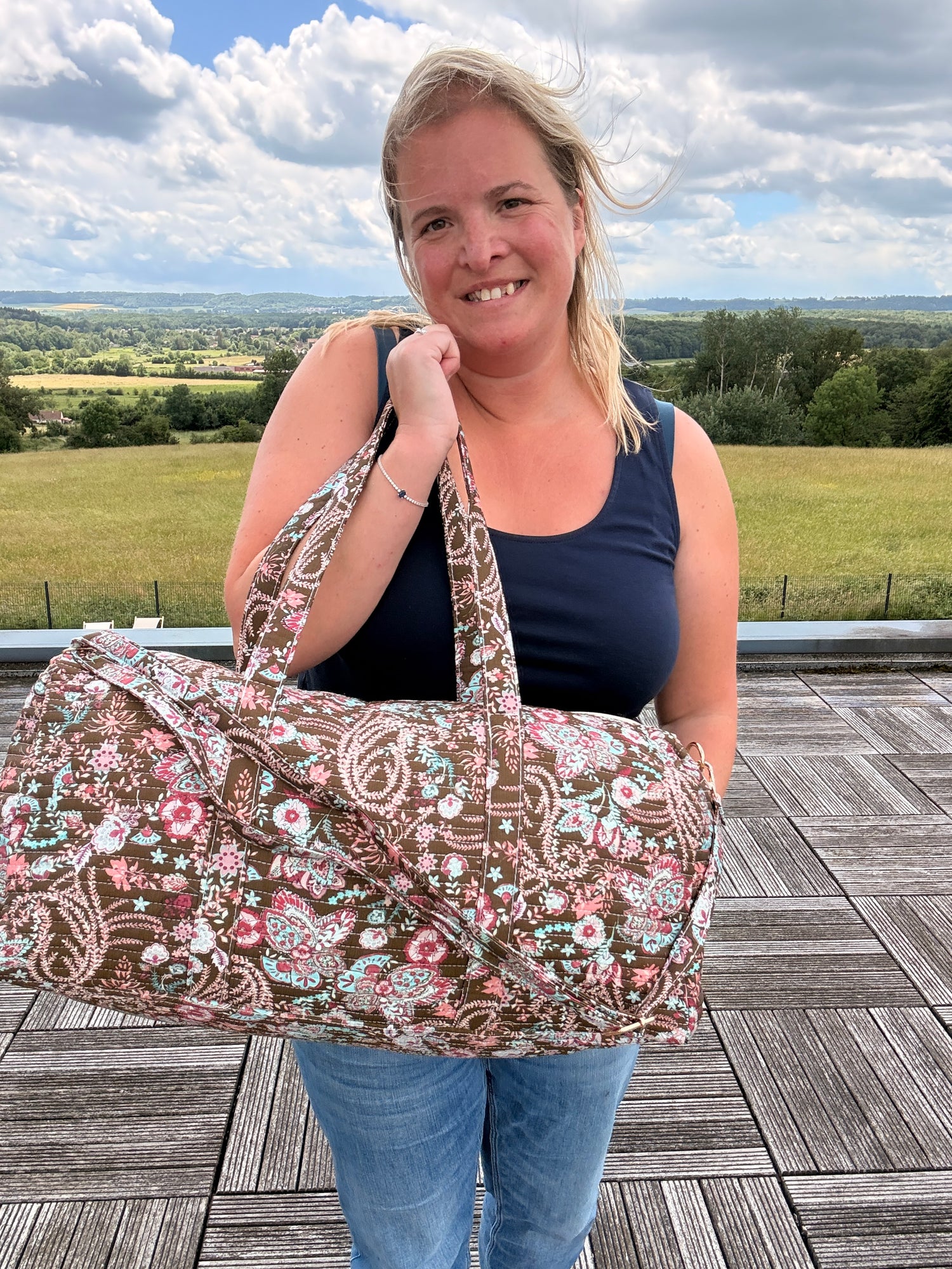 Sac polochon matelassé fleuri Paisley porté à l’épaule par une femme Souriante, grand sac de voyage bohème chic avec bandoulière incluse.