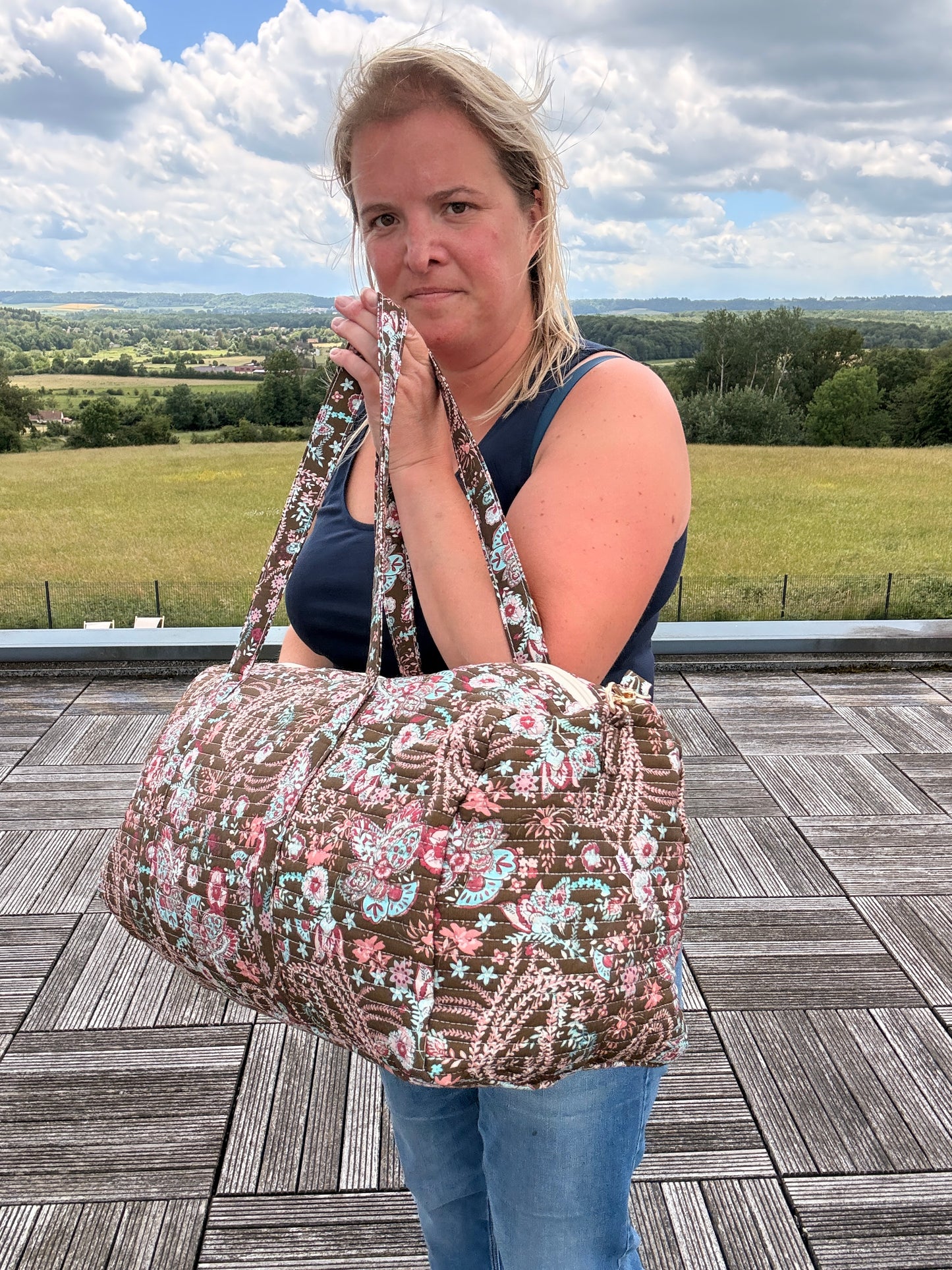 Sac polochon matelassé fleuri paisley, grand sac de voyage boho chic, femme porte-bandoulière incluse sur fond paysage.