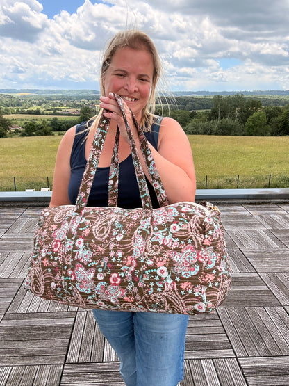 Sac polochon matelassé fleuri Paisley, grande bandoulière incluse, femme bohème tenant le sac de voyage sur fond terrasse et paysage.