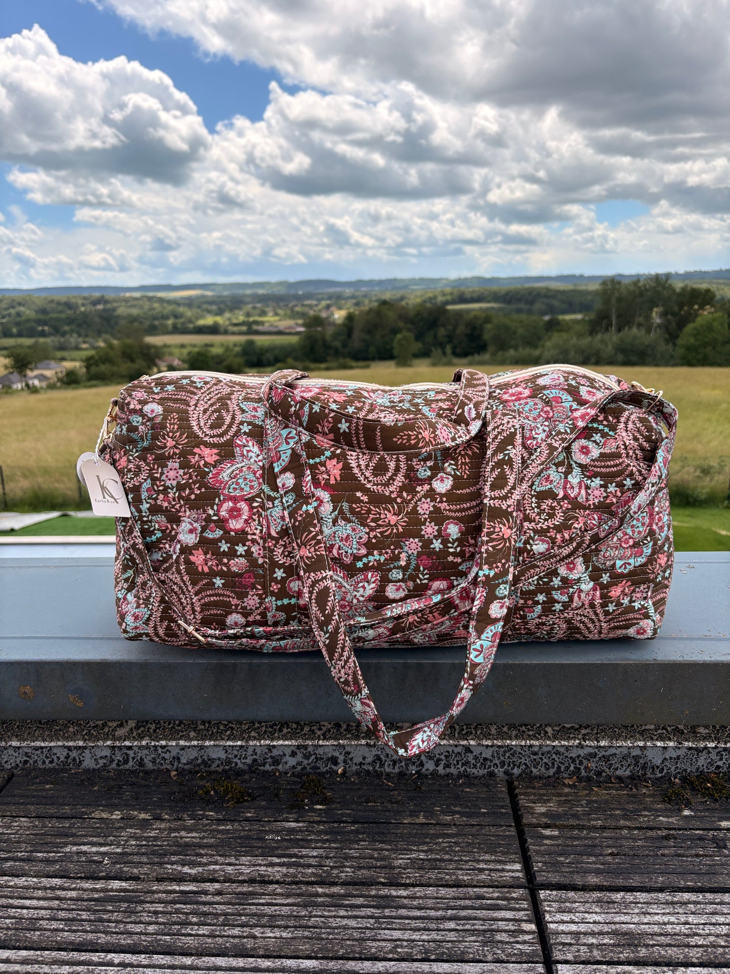 Sac polochon matelassé fleuri Paisley posé en extérieur, bandoulière incluse, motif bohème chic sur fond marron.