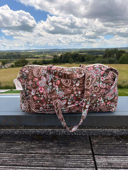 Sac polochon matelassé fleuri Paisley posé en extérieur, bandoulière incluse, motif bohème chic sur fond marron.