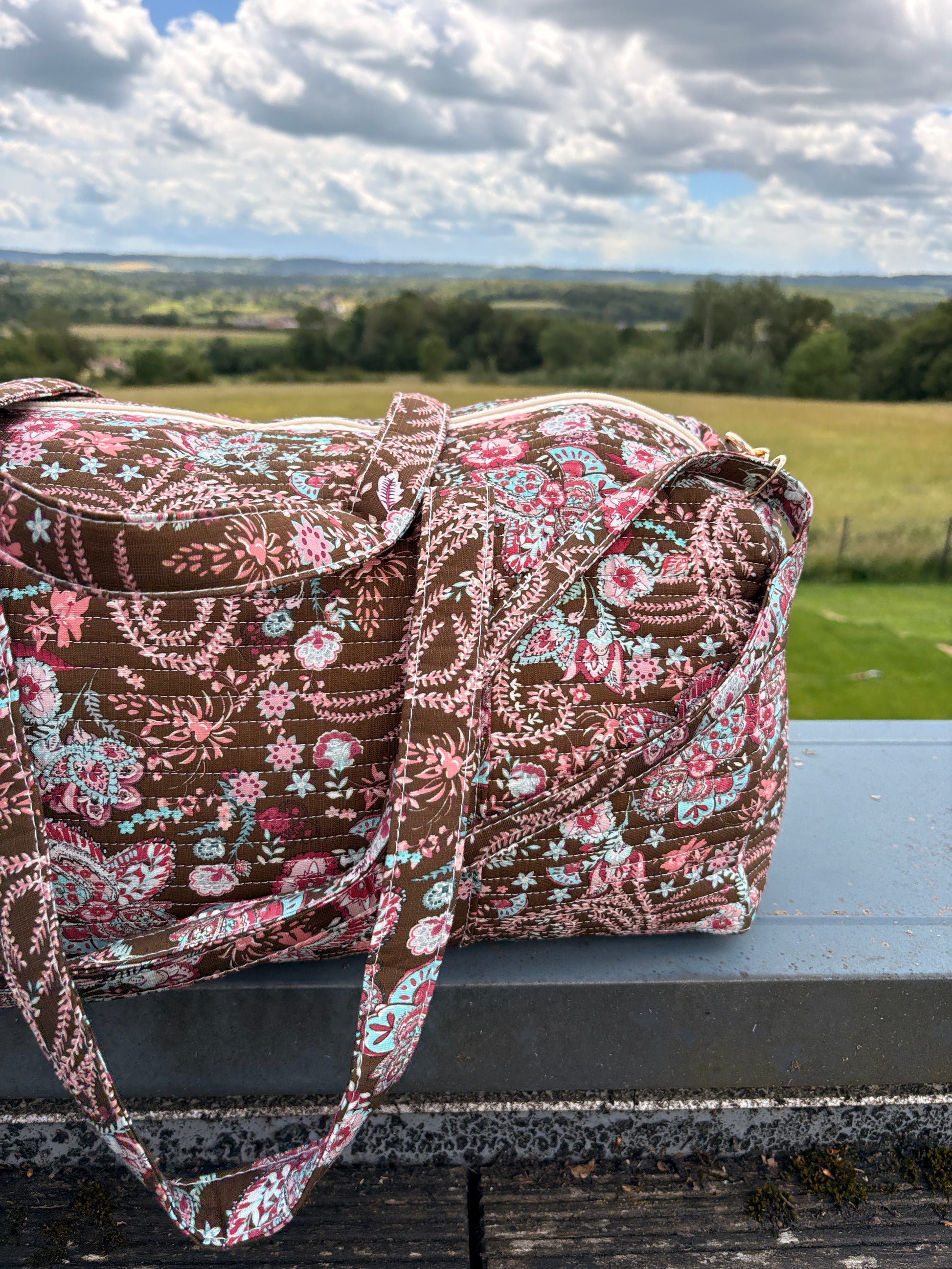 Sac polochon matelassé fleuri paisley posé en extérieur, bandoulière incluse et motif brun, rose et bleu pastel.