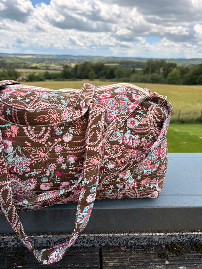 Sac polochon matelassé fleuri paisley posé en extérieur, bandoulière incluse et motif brun, rose et bleu pastel.