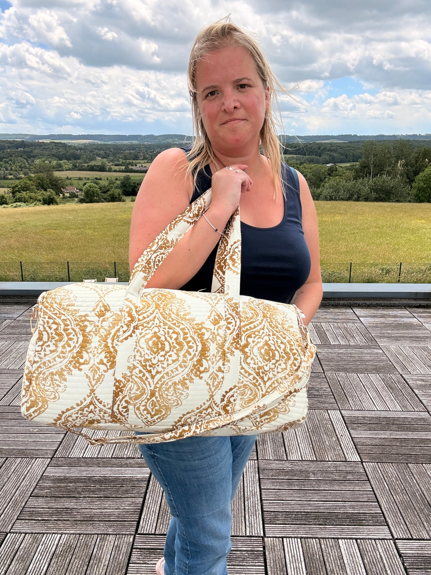 Sac polochon matelassé imprimé baroque doré porté par une jeune femme sur une terrasse, paysage champêtre en arrière-plan.