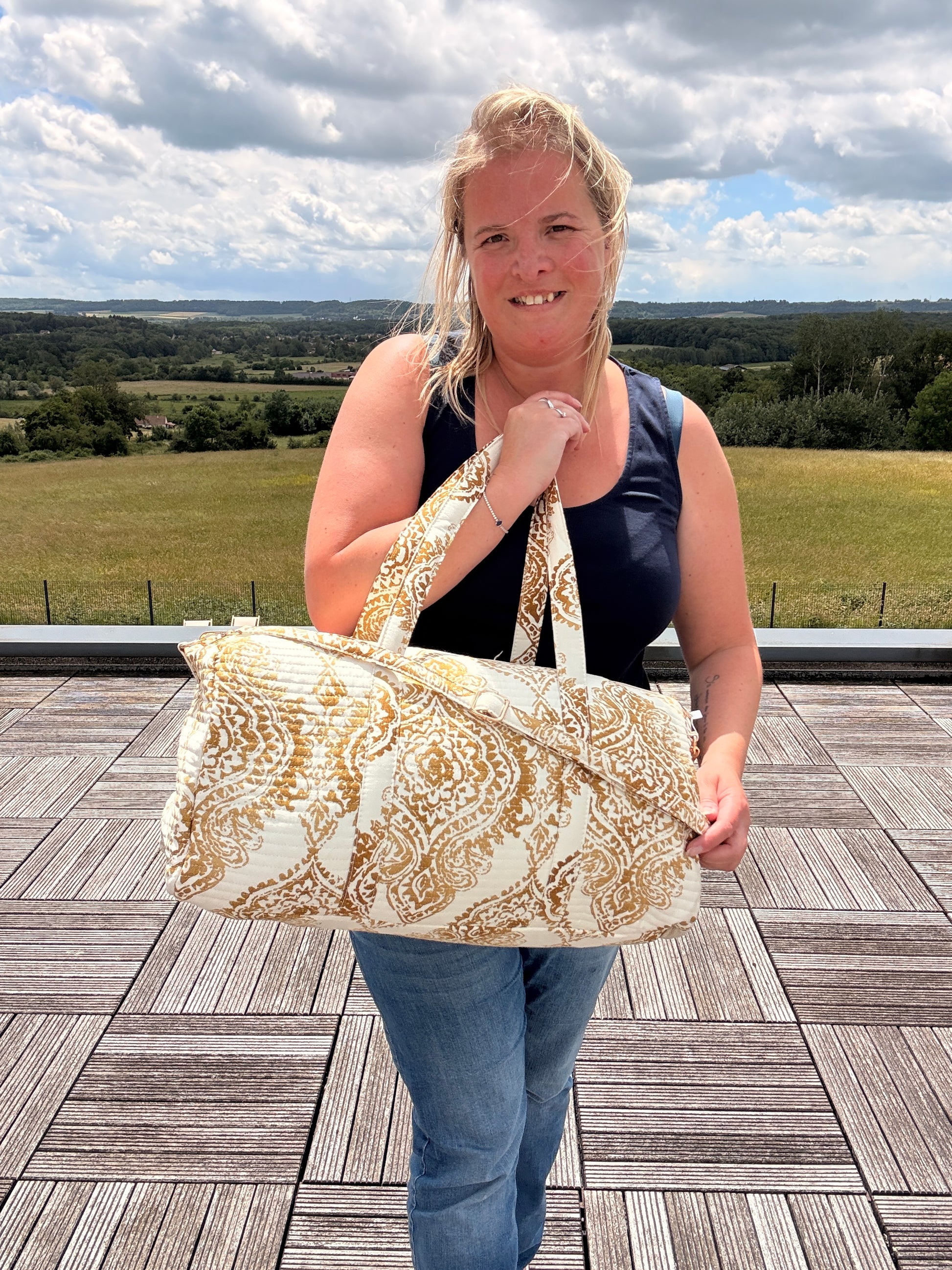 Sac polochon matelassé imprimé baroque doré porté par une femme sur une terrasse, paysage rural en arrière-plan.