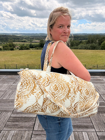 Sac polochon matelassé imprimé baroque doré porté sur l’épaule d’une femme, paysage champêtre en arrière-plan.