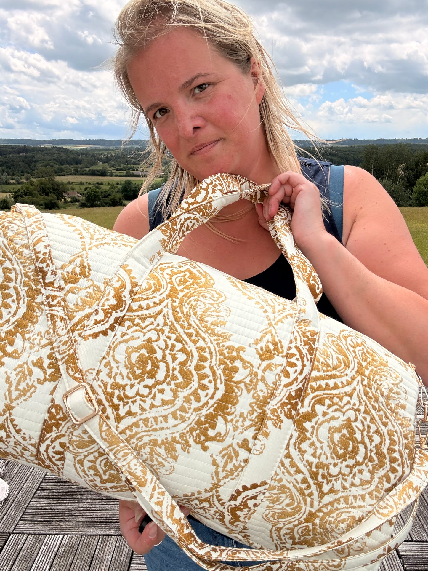 Sac polochon matelassé imprimé baroque doré porté par une femme, décor naturel en arrière-plan.