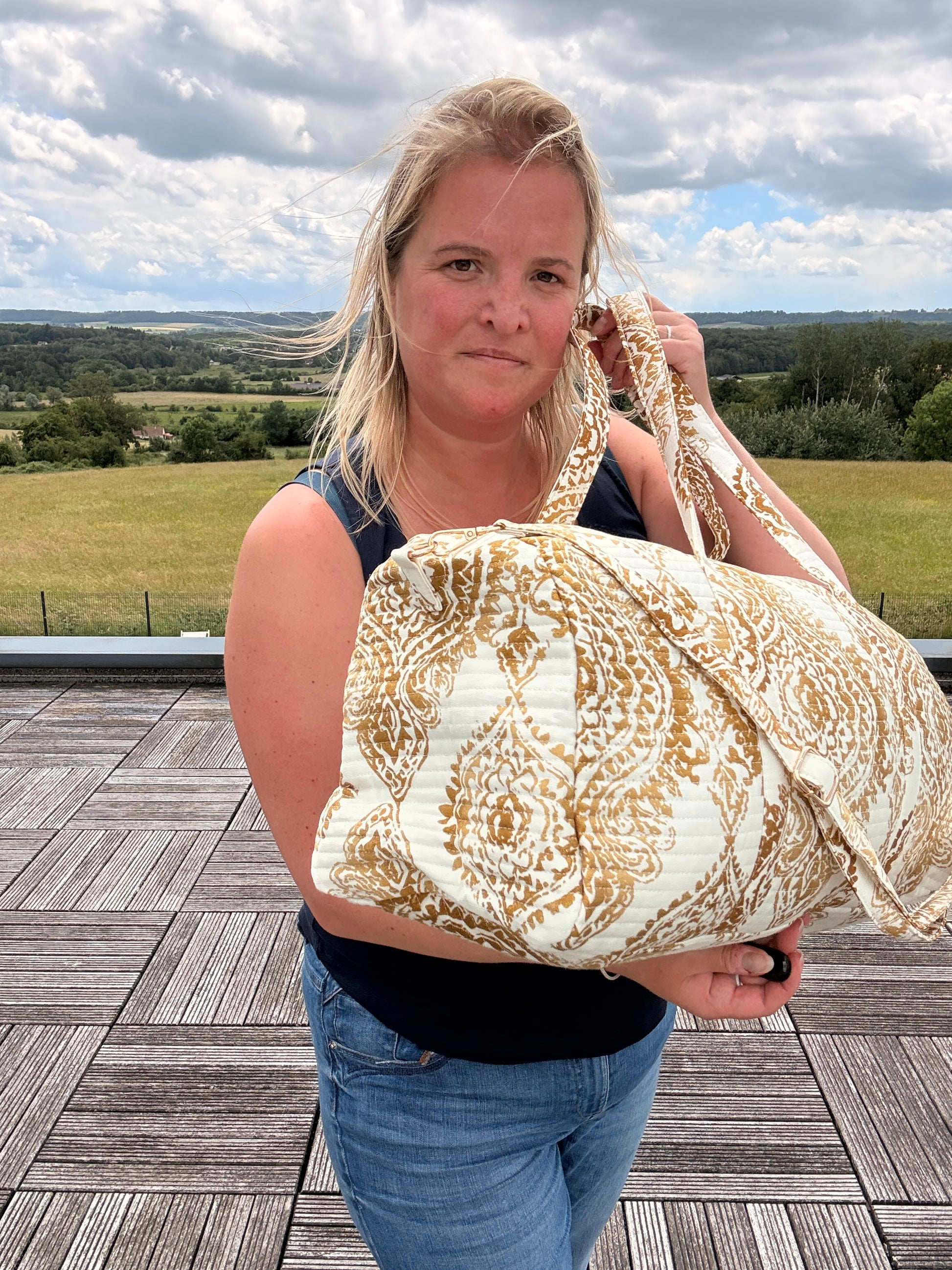 Femme tenant un sac polochon matelassé imprimé baroque doré sur fond clair, en plein air sur une terrasse, paysage champêtre en arrière-plan. Sac voyage femme bohème chic.
