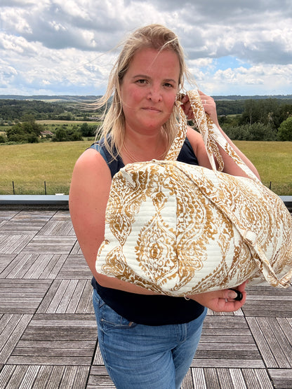 Femme tenant un sac polochon matelassé imprimé baroque doré sur fond clair, en plein air sur une terrasse, paysage champêtre en arrière-plan. Sac voyage femme bohème chic.