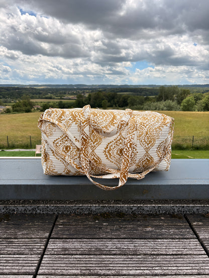 Sac polochon matelassé imprimé baroque doré posé sur une rambarde avec paysage champêtre en arrière-plan. Sac voyage femme bohème chic.