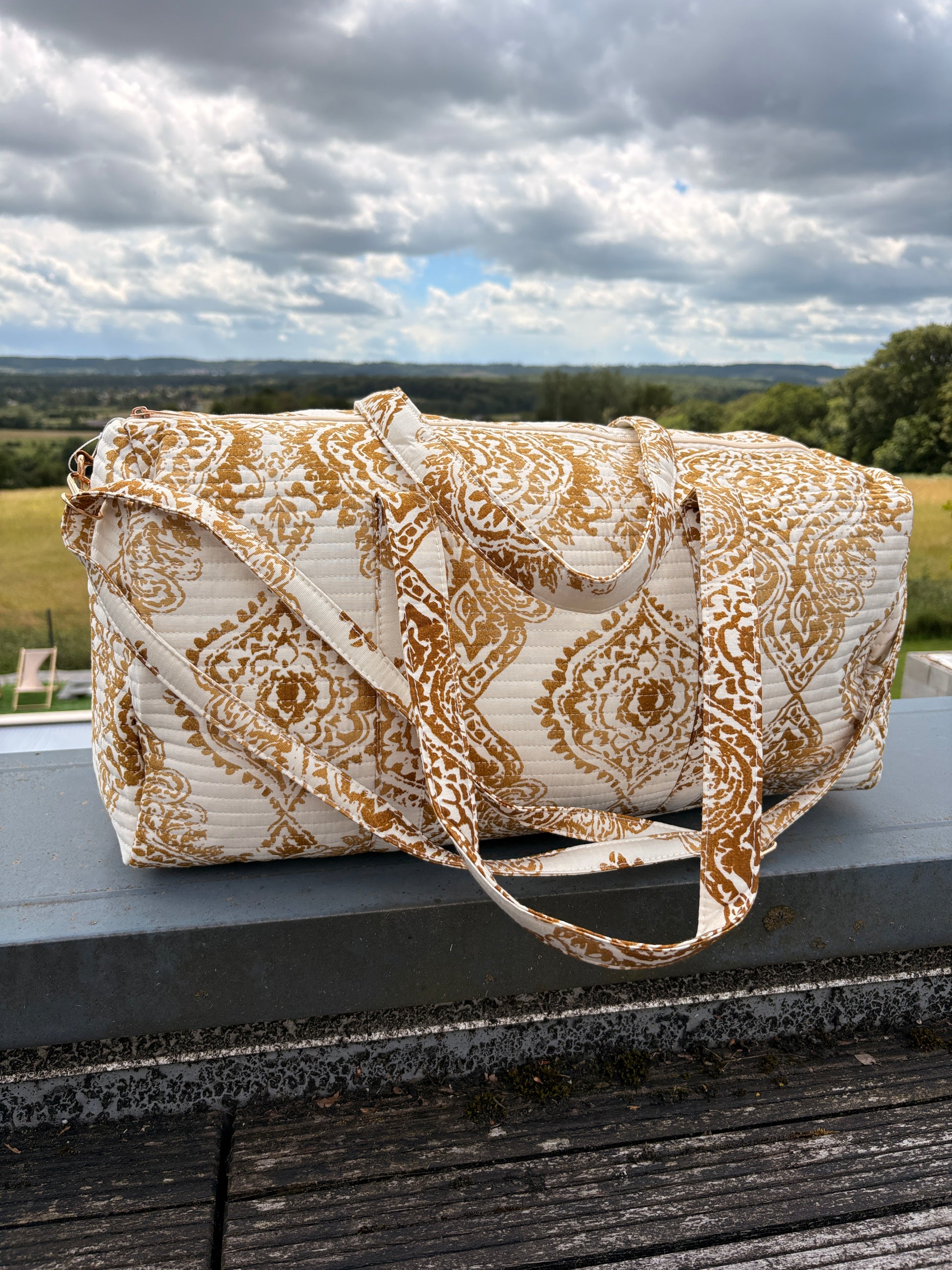 Sac polochon matelassé imprimé baroque doré posé sur une rambarde, motif bohème chic et bords de paysage en arrière-plan.