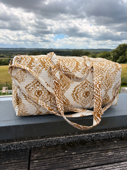 Sac polochon matelassé imprimé baroque doré posé sur une rambarde, motif bohème chic et bords de paysage en arrière-plan.