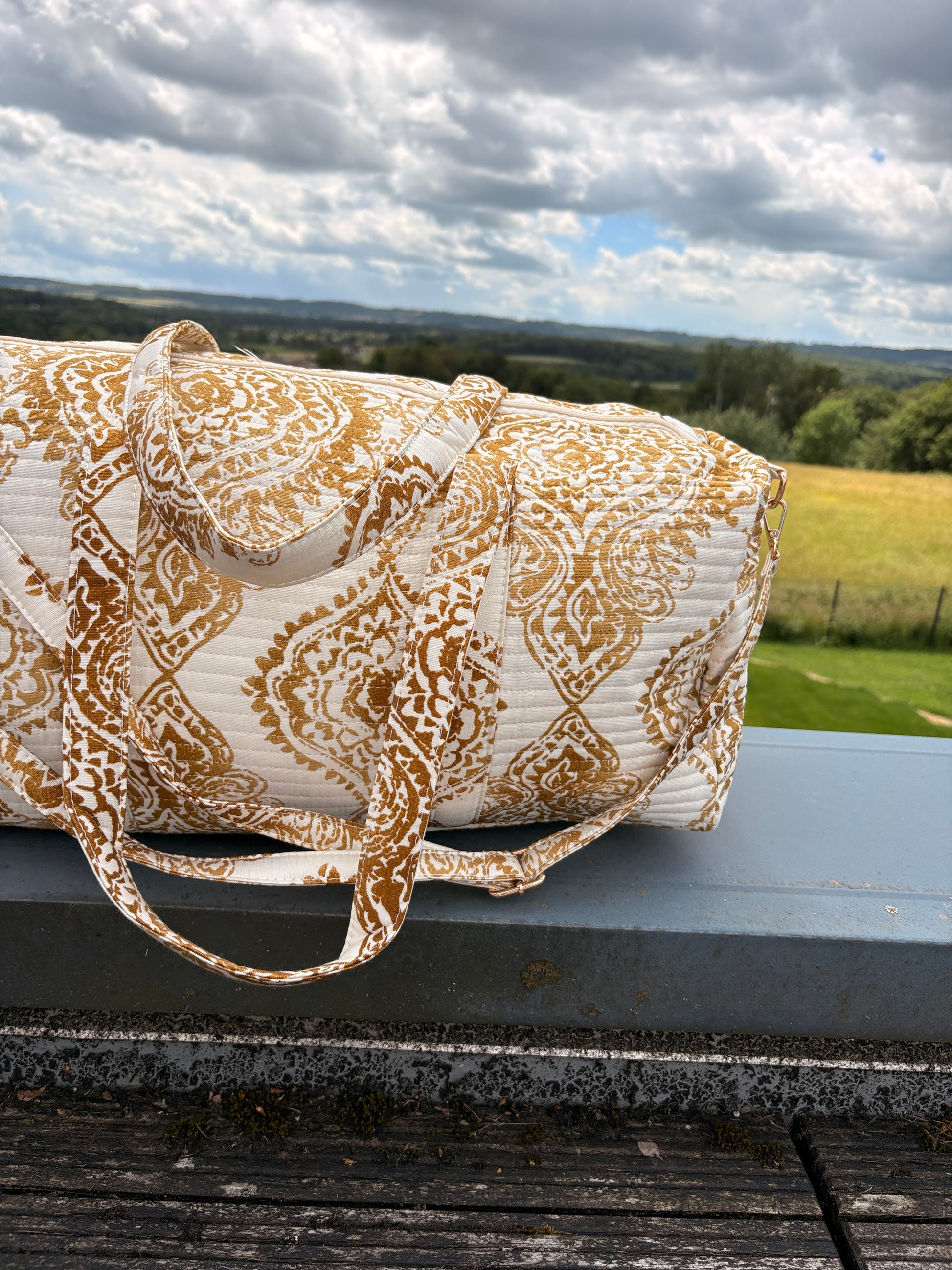 Sac polochon matelassé imprimé baroque doré posé sur une surface extérieure, avec poignées assorties et paysage champêtre en arrière-plan.