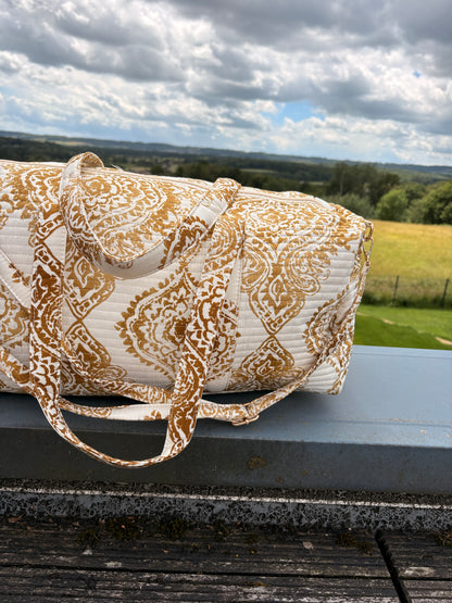 Sac polochon matelassé imprimé baroque doré posé sur une surface extérieure, avec poignées assorties et paysage champêtre en arrière-plan.