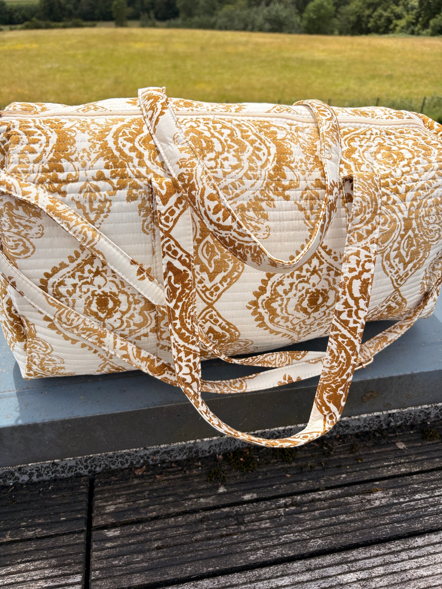 Sac polochon matelassé imprimé baroque doré, bagage voyage femme bohème chic sur plateau en bois.