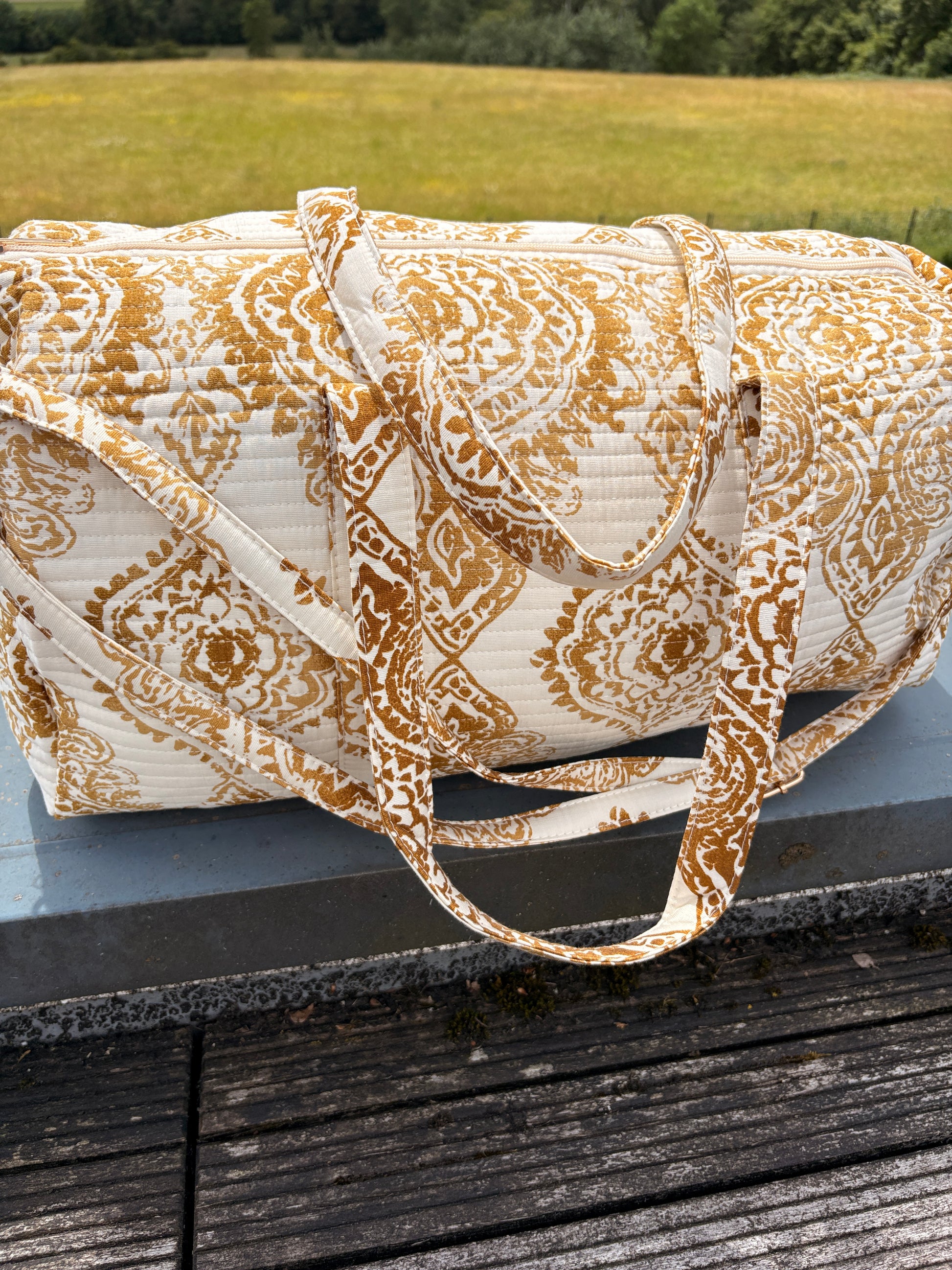 Sac polochon matelassé imprimé baroque doré, bagage voyage femme bohème chic sur plateau en bois.