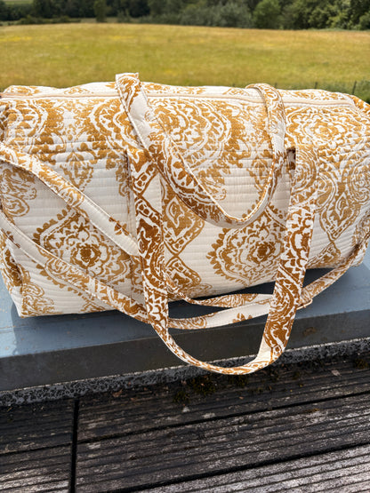 Sac polochon matelassé imprimé baroque doré, bagage voyage femme bohème chic sur plateau en bois.