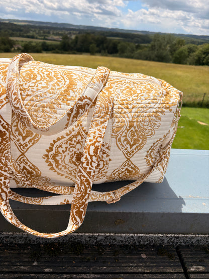 Sac polochon matelassé imprimé Baroque doré posé sur une rambarde, avec sa lanière assortie, en paysage champêtre.