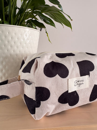 Grande trousse maquillage femme motifs cœurs noirs, zippe en tissu léger, avec étiquette “Boutique Bazar” sur fond beige, près d’une plante.