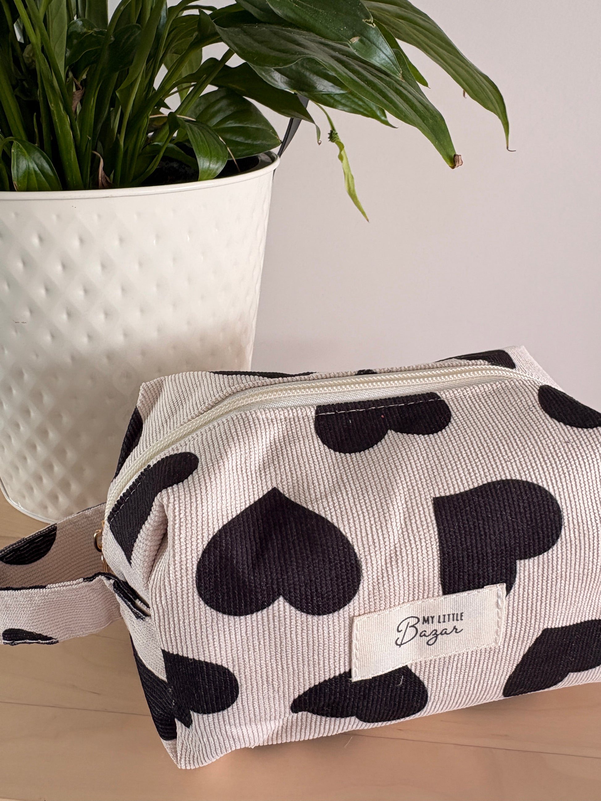 Pochette cosmétique zippée à motif coeurs noirs sur fond clair, près d’un plant en pot blanc. Porte-étiquette "My Little Bag" sur le devant.