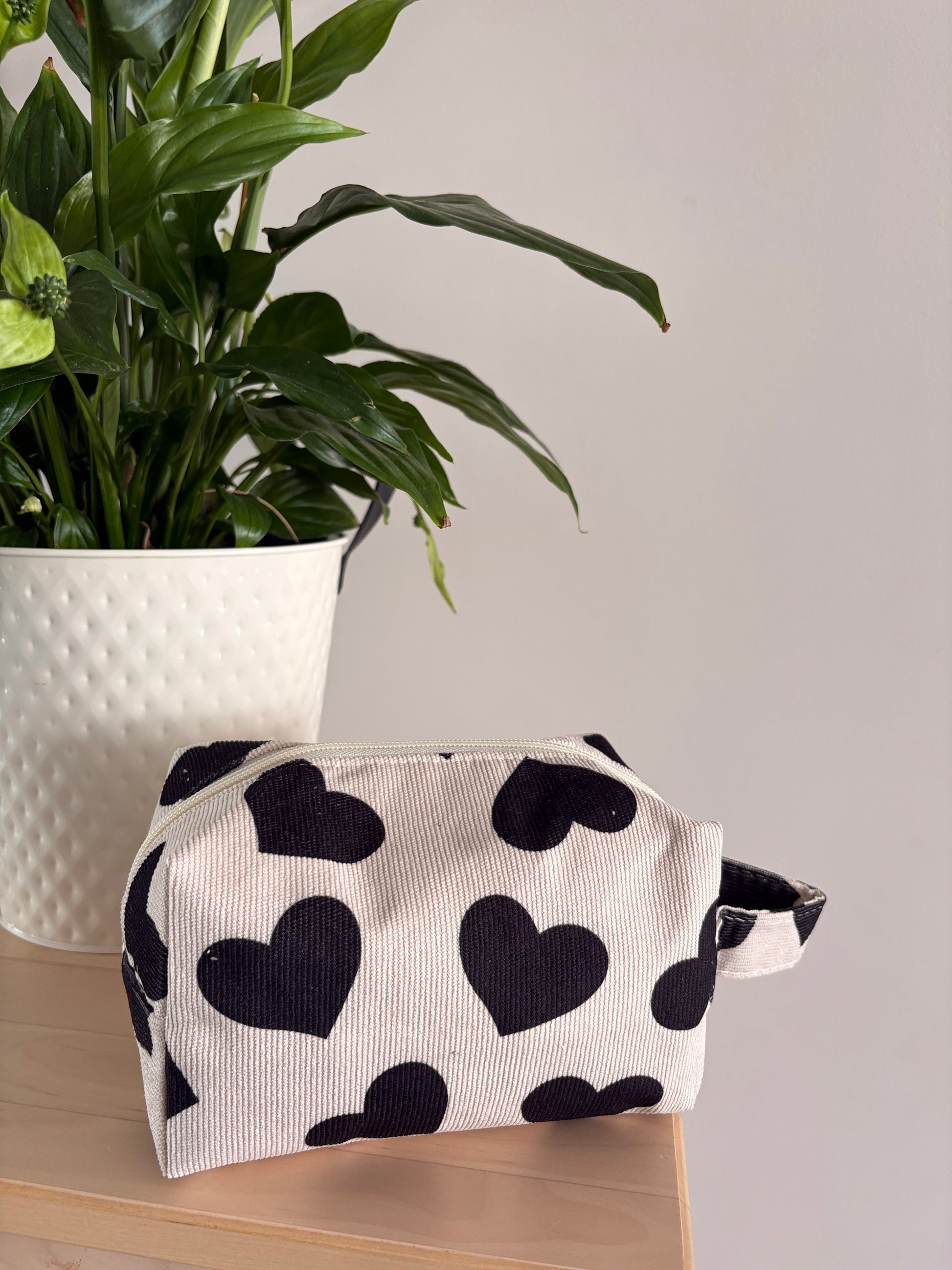 Petite trousse maquillage à motif cœurs noirs sur fond blanc, zippée, posée sur une table près d’une plante.