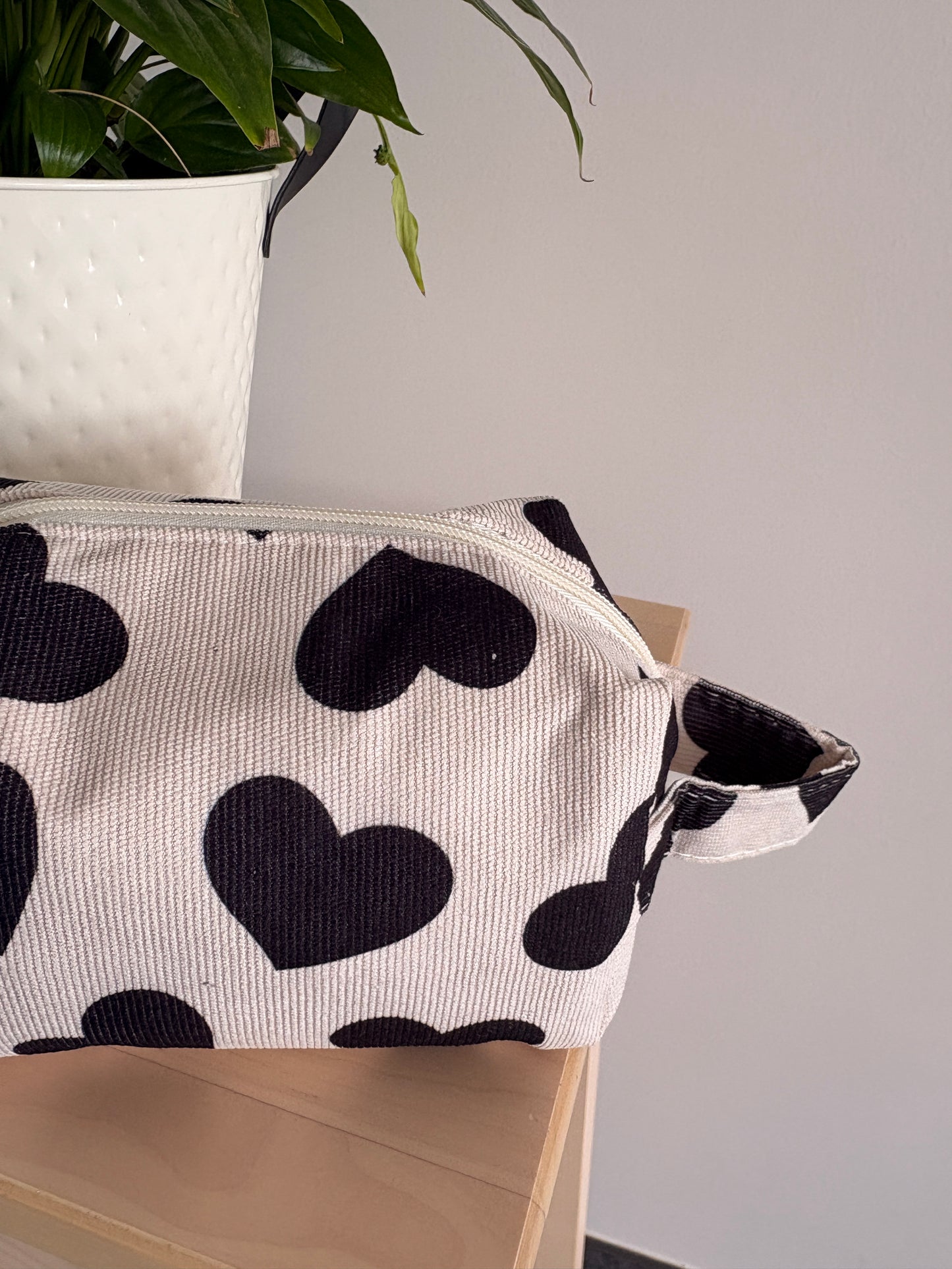 Grande trousse maquillage femme motifs cœurs noirs sur tissu clair, zippée, poignée latérale, posé sur meuble.
