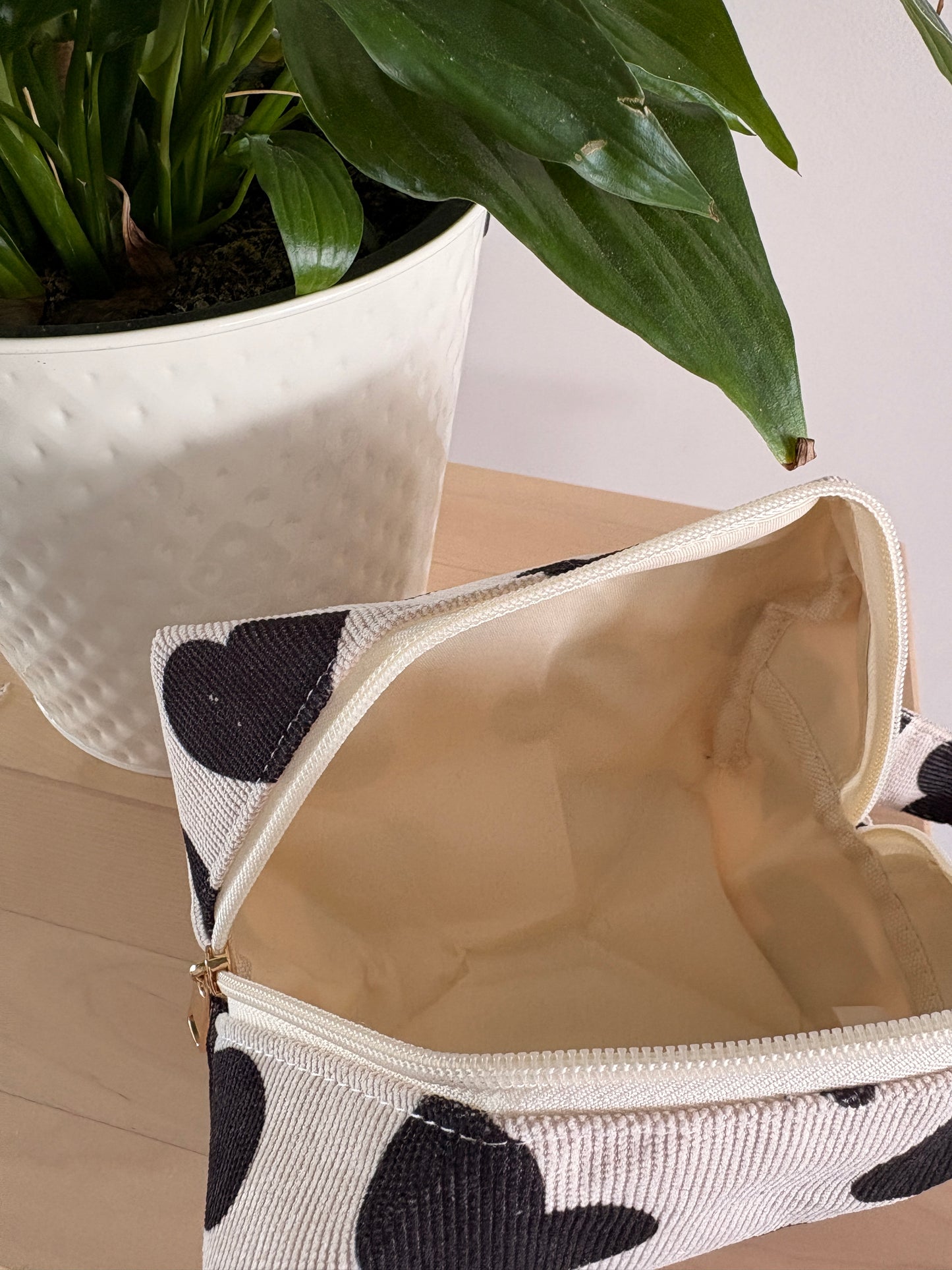Grande trousse maquillage féminine motif cœurs noirs, zippée, ouverte sur table avec plante en pot à côté.