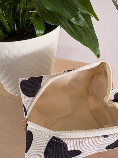 Grande trousse maquillage féminine motif cœurs noirs, zippée, ouverte sur table avec plante en pot à côté.