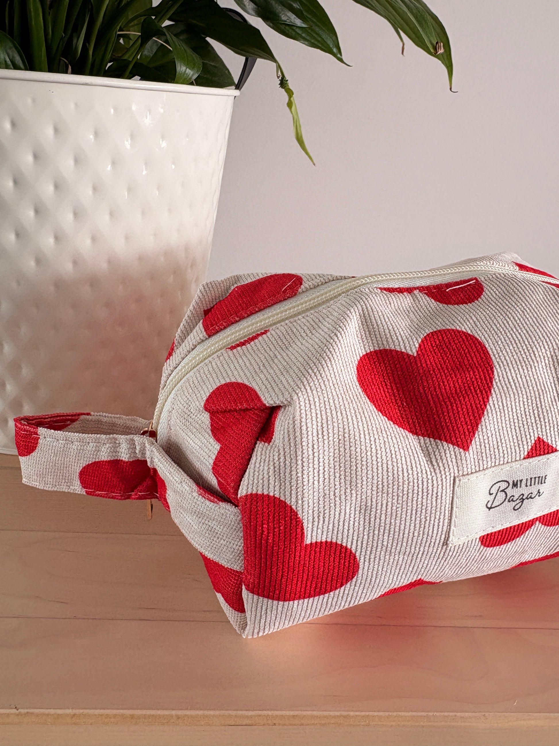 Trousse de maquillage pour femme motif cœurs, pochette cosmétique zippée sur fond beige, près d’un pot décoratif.