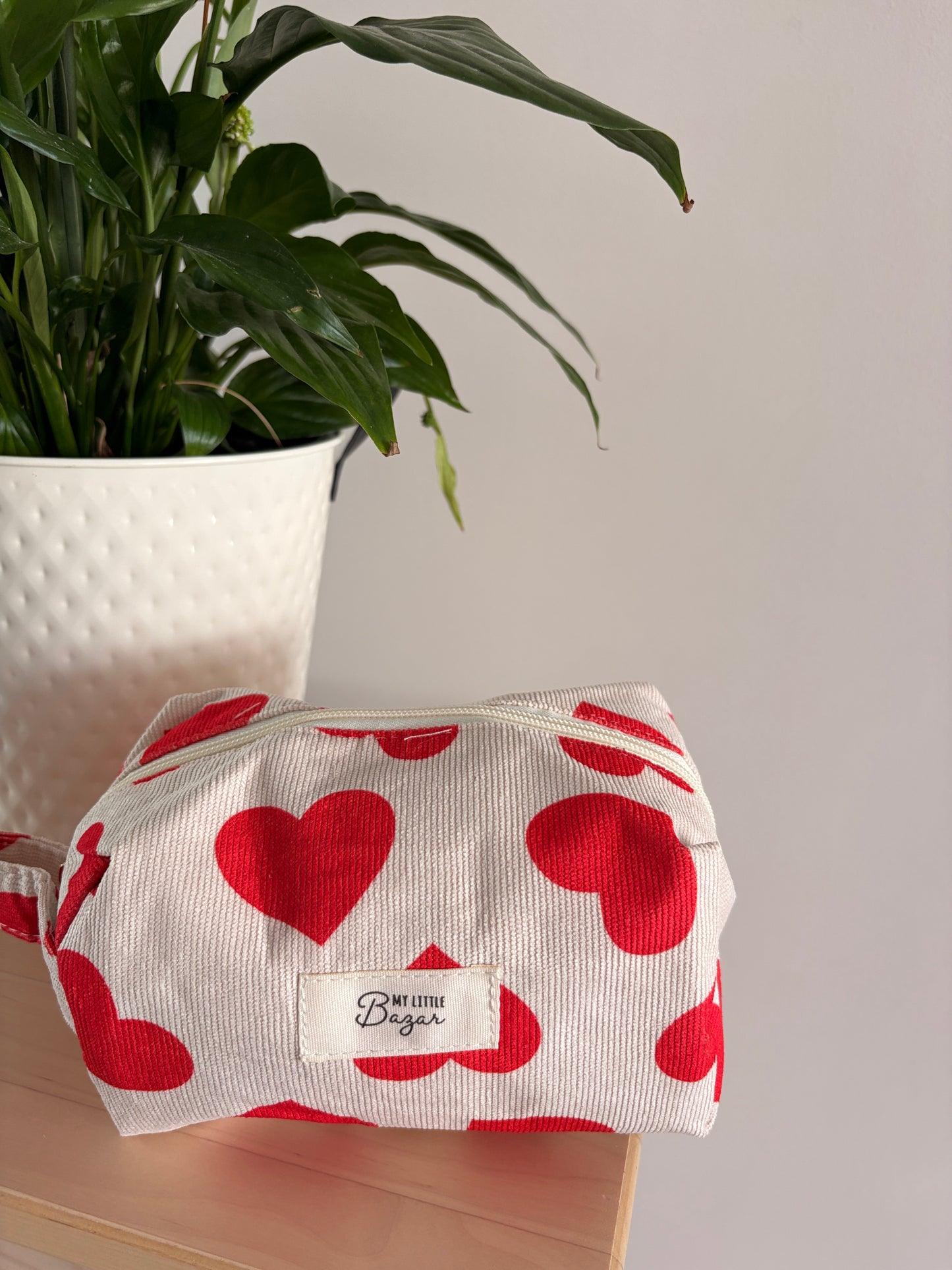 Trousse de Maquillage Femme Motif Cœurs sur fond clair avec zippette, près d’une plante en pot.