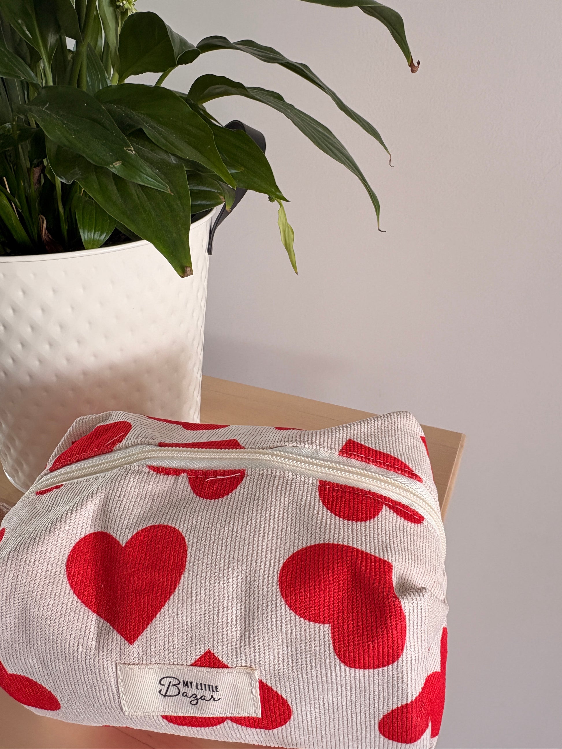 Trousse de maquillage femme motif cœurs blanche avec zips rouges, posée sur une table près d’une plante décorative.
