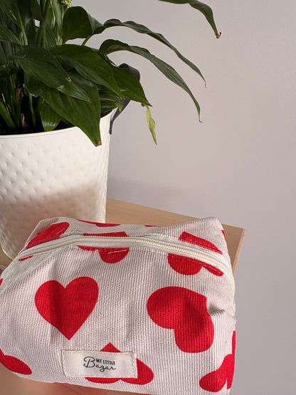 Trousse de maquillage femme motif cœurs blanche avec zips rouges, posée sur une table près d’une plante décorative.