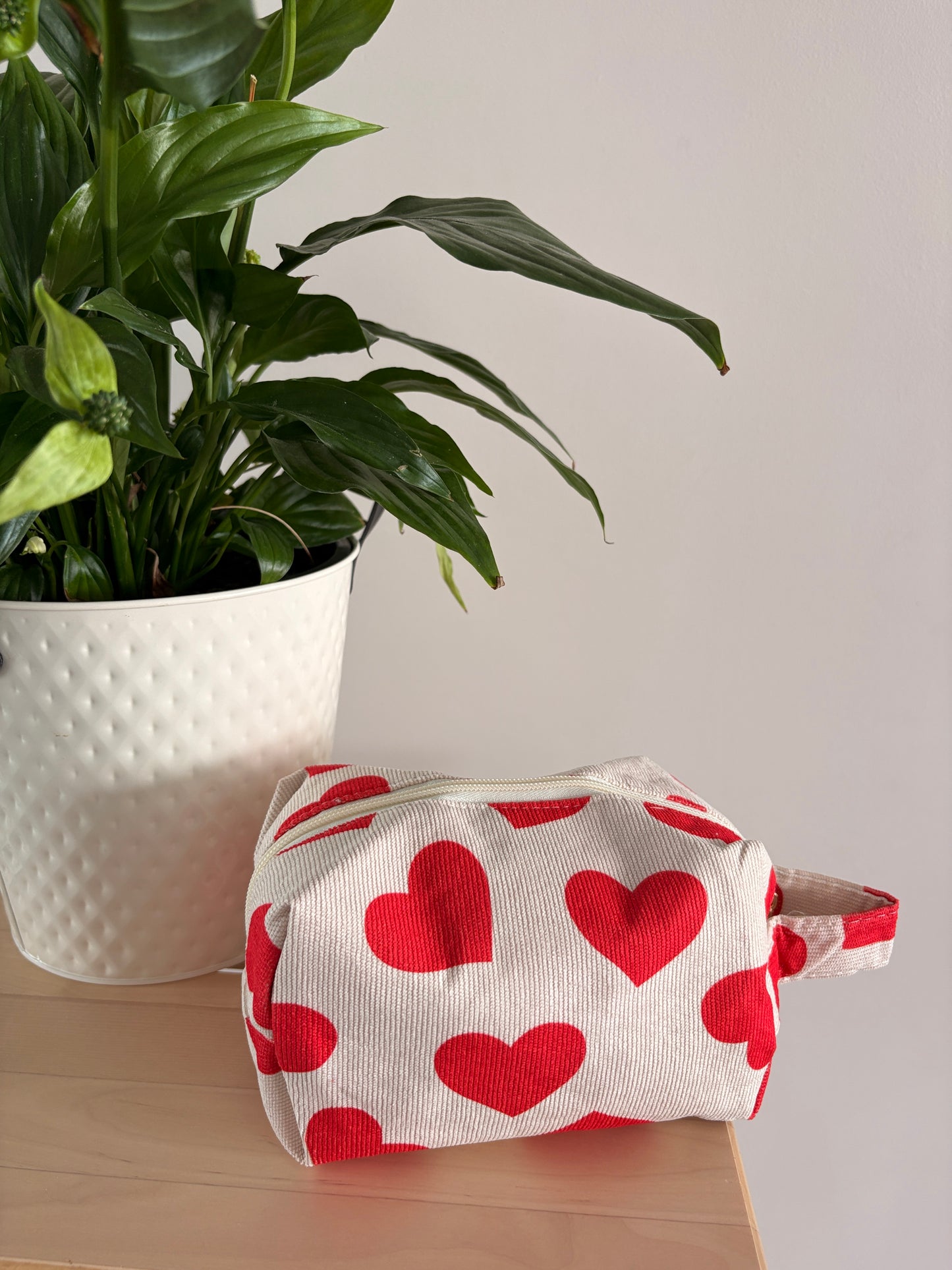 Trousse de maquillage avec motif cœurs rouges sur fond blanc, pochette zippée posée près d’une plante verte.