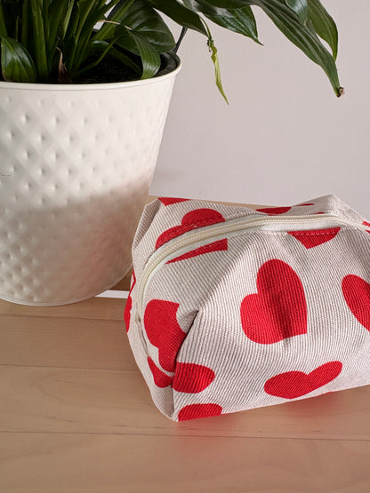 Trousse de maquillage féminine à motif cœurs rouges sur fond clair, zippée, posée à côté d’un pot de plante sur une surface en bois.