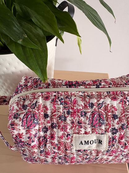 Grande trousse maquillage imprimé floral rose avec étiquette AMOUR, pochettes zippées et grande capacité, posée près d’une plante.