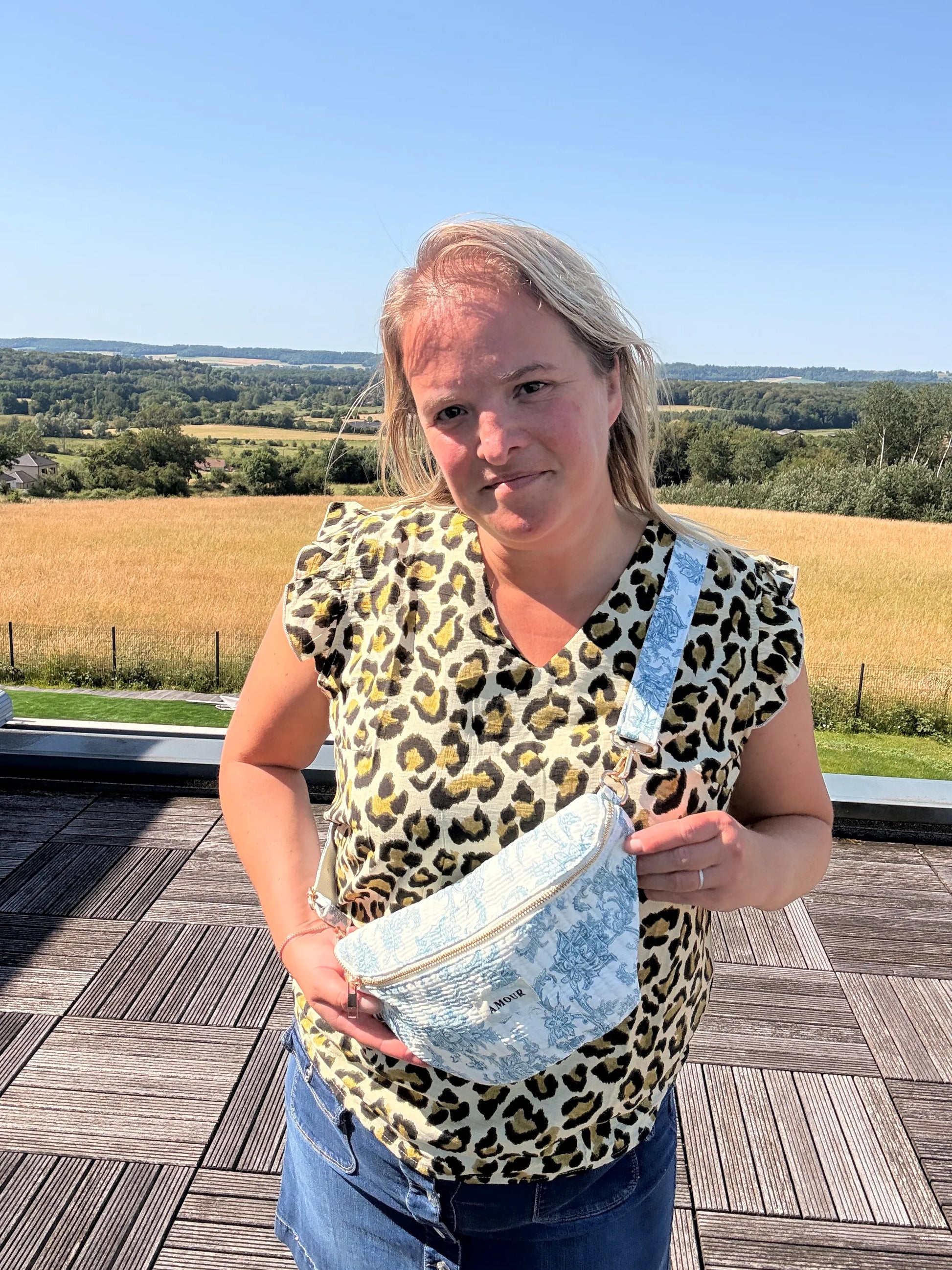 Sac banane Toile de Jouy – Imprimé bleu turquoise porté en bandoulière par une femme sur une terrasse, paysage champêtre en arrière-plan.