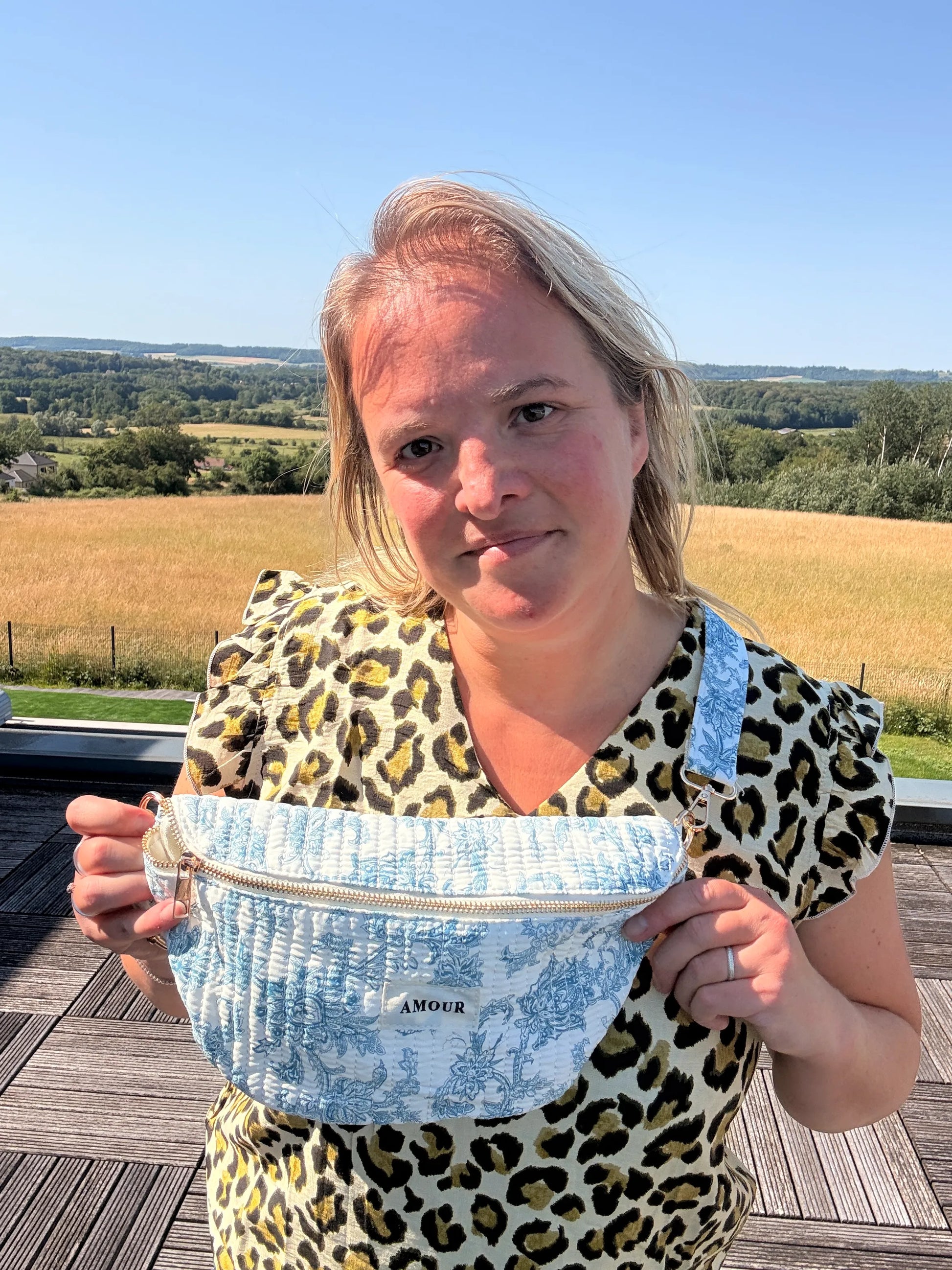 Sac Banane Toile de Jouy – Imprimé Bleu Turquoise porté en bandoulière sur l’épaule, femme debout sur une terrasse avec terrain et campagne en arrière-plan. Amour imprimé sur la poche.