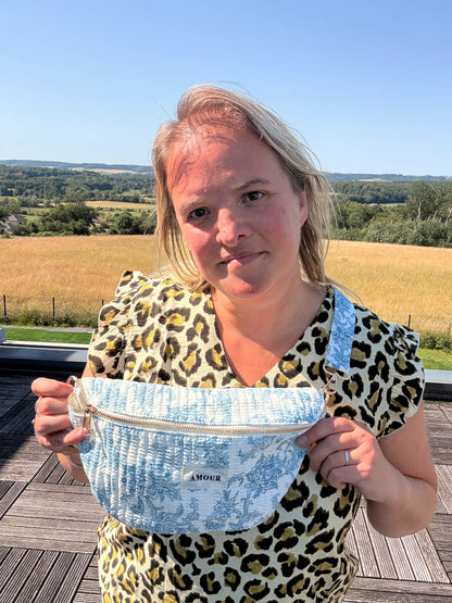 Sac Banane Toile de Jouy – Imprimé Bleu Turquoise porté en bandoulière sur l’épaule, femme debout sur une terrasse avec terrain et campagne en arrière-plan. Amour imprimé sur la poche.